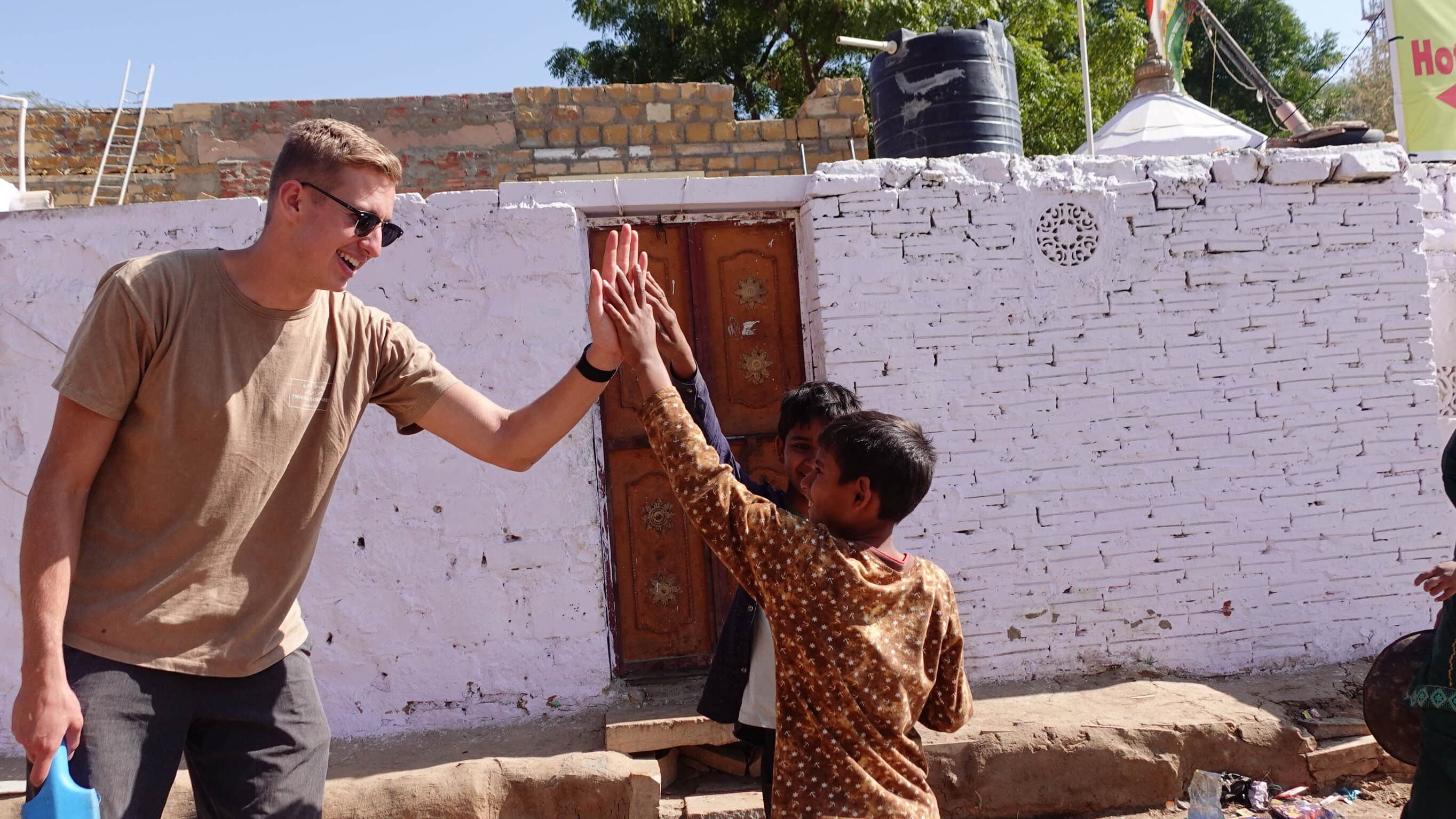 A smiling Western man in sunglasses gives a high-five to local children in front of a whitewashed brick wall in a South Asian village
