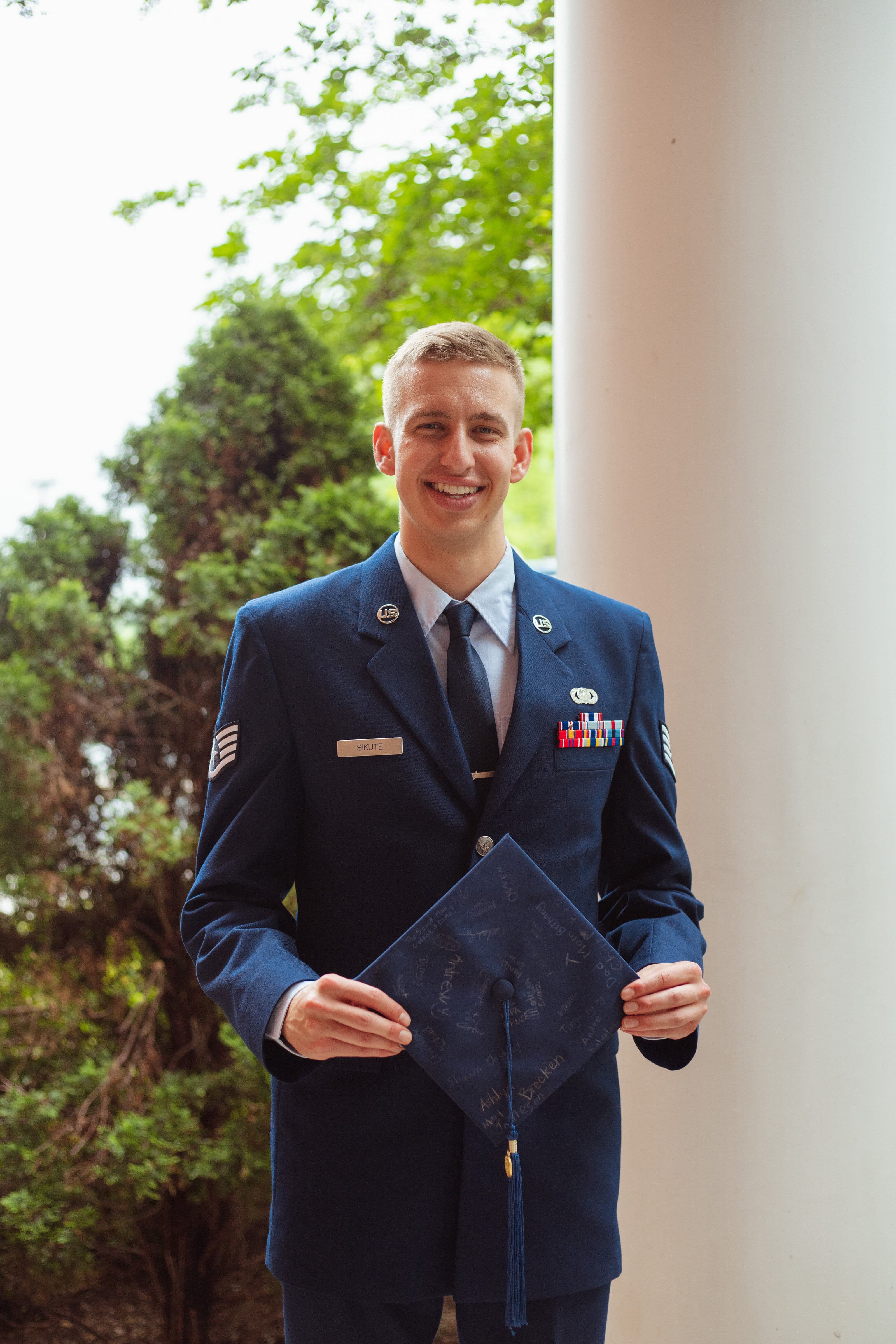 Smiling US Air Force servicemember in dress blues holding a signed graduation cap while standing beside a white column outdoors