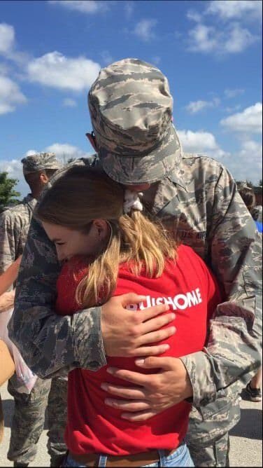 A US Air Force service member in camouflage uniform embraces a young girl in a red shirt outdoors at a military gathering