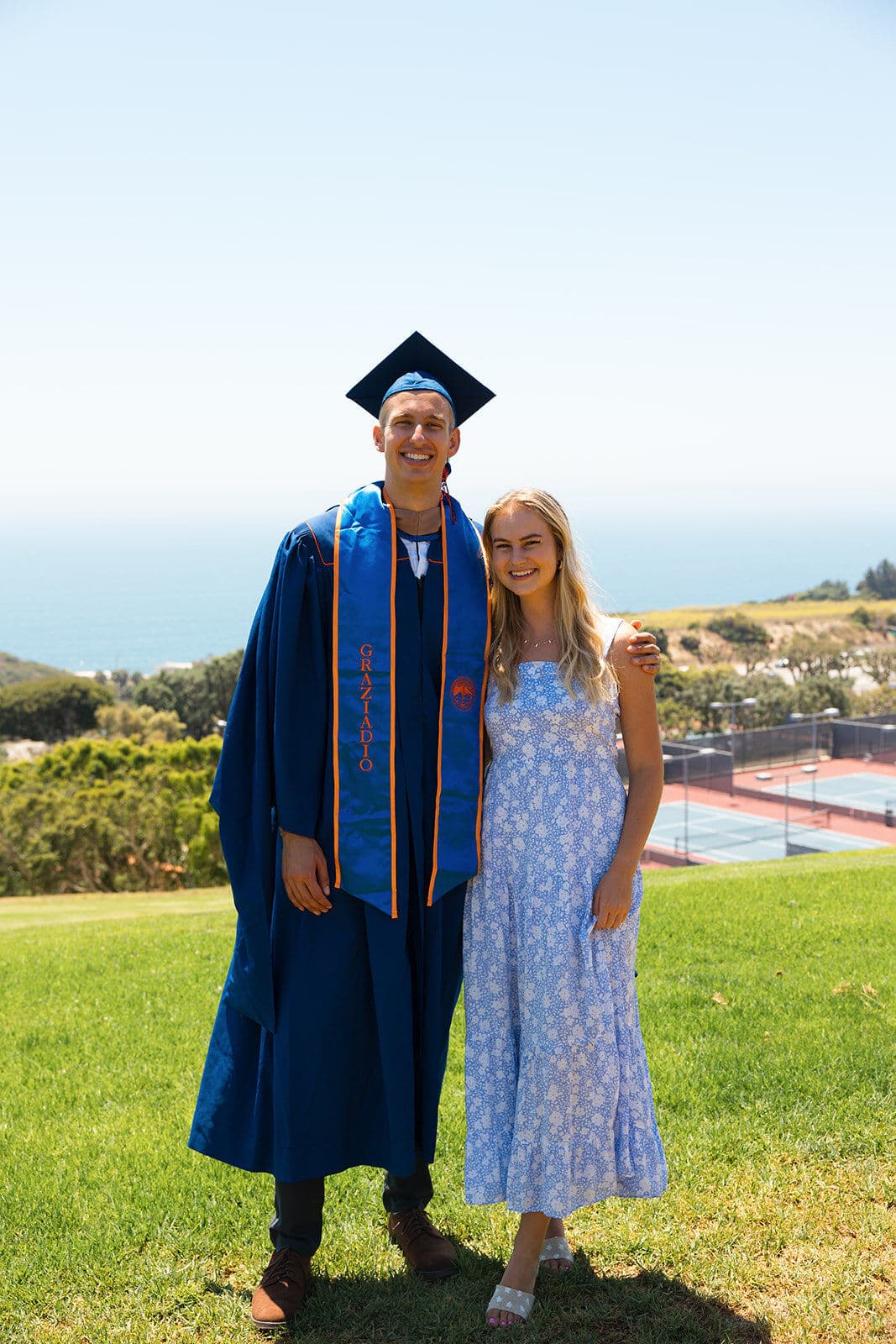 A tall male graduate in a blue cap and gown with a Graziadio stole stands with a young woman in a floral dress on a green hillside overlooking the ocean and tennis courts