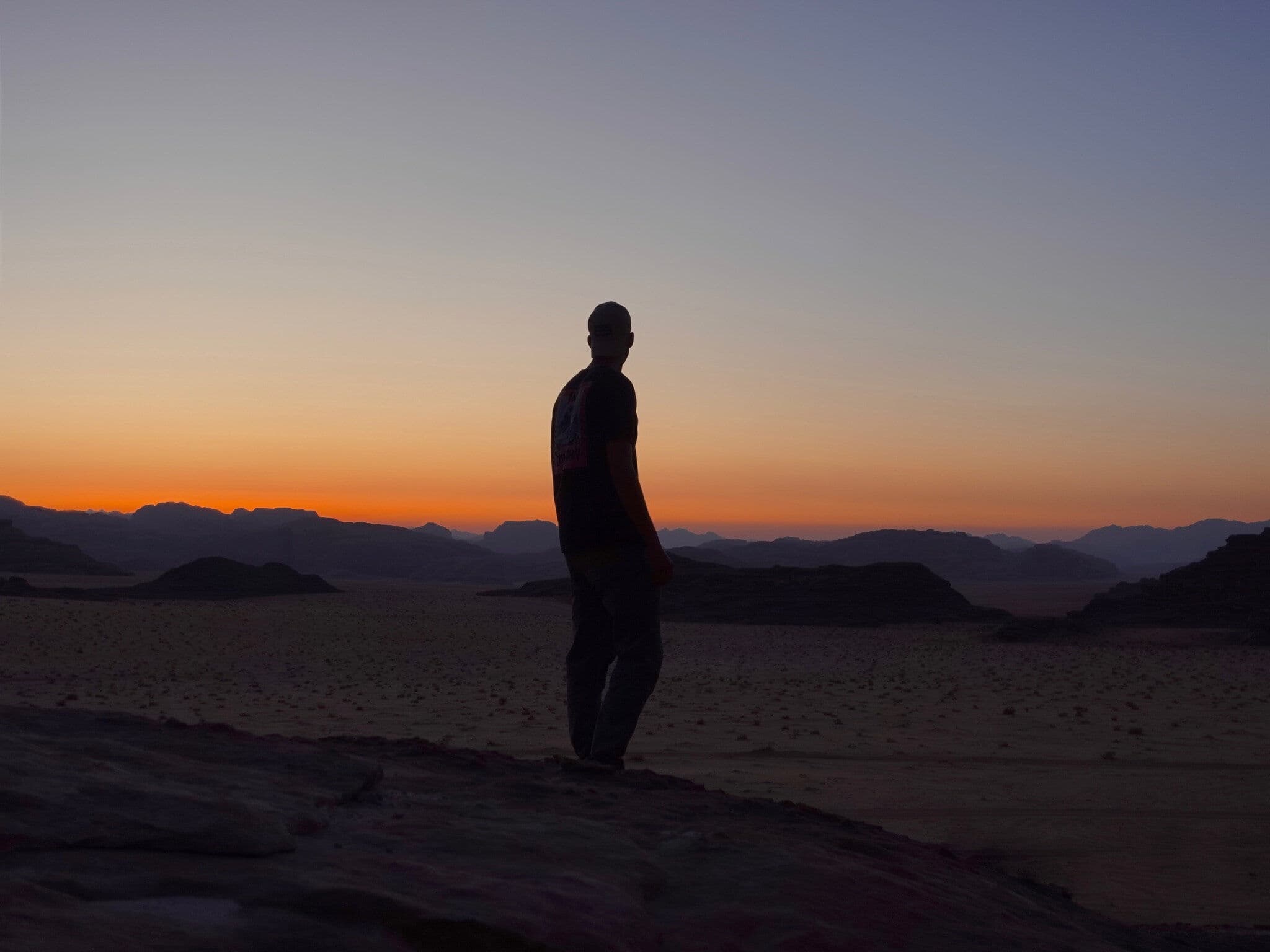 Silhouette of a person standing on a rocky desert landscape at sunset, with layered mountain ridges and a vivid orange horizon in the background