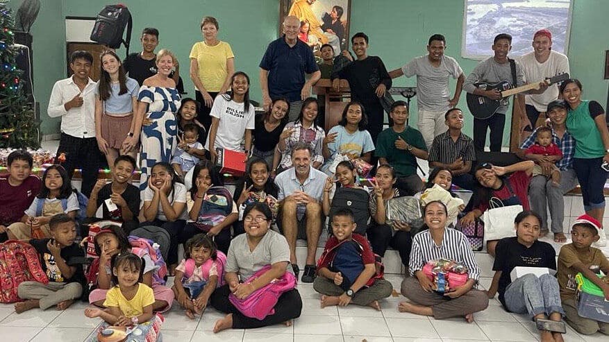Large group of adults and children posing together indoors with gifts and backpacks, with a religious painting visible on the wall behind them