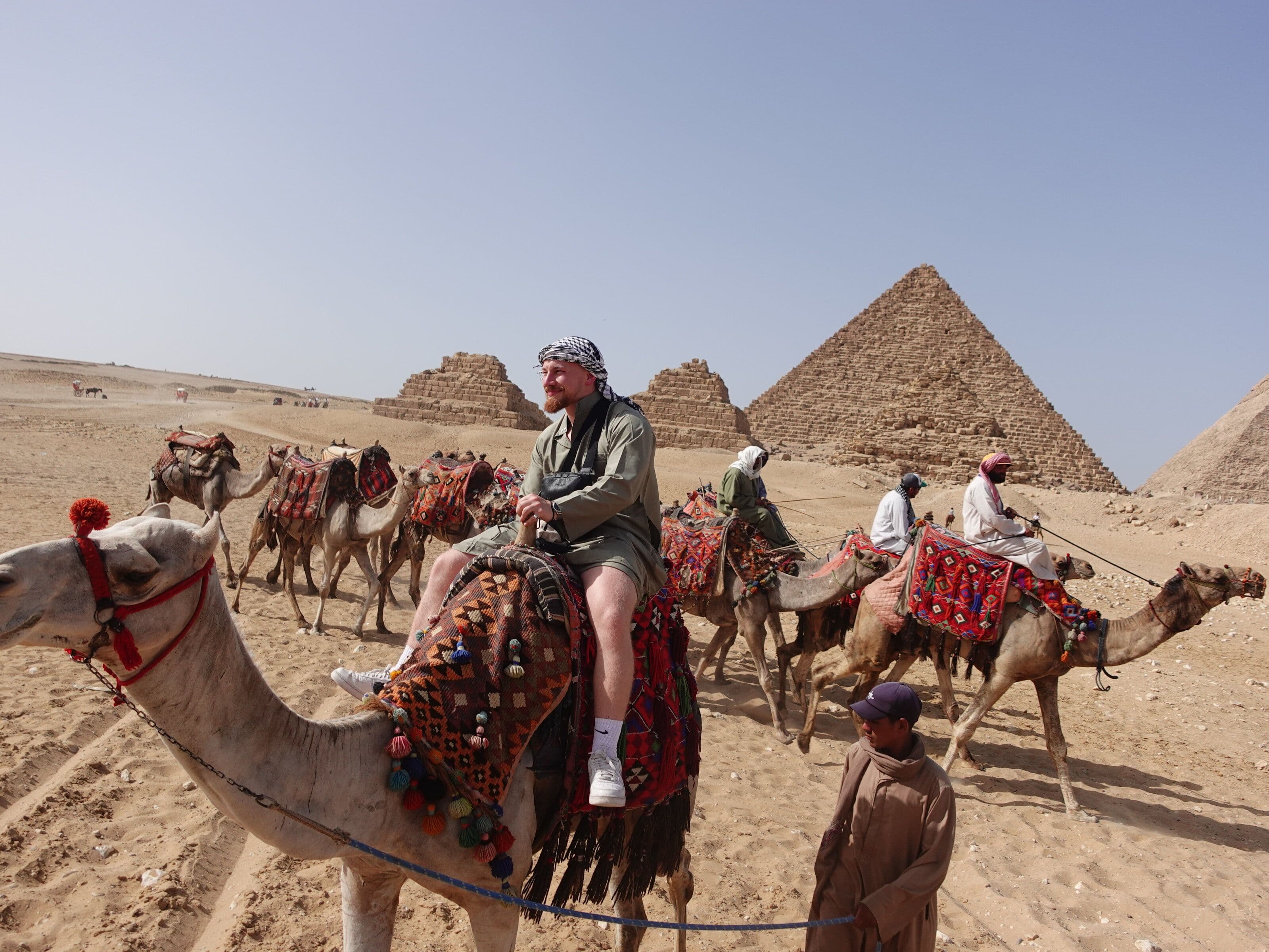 Red-haired man riding a decorated camel in front of the Pyramids of Giza, Egypt, with a camel handler standing nearby