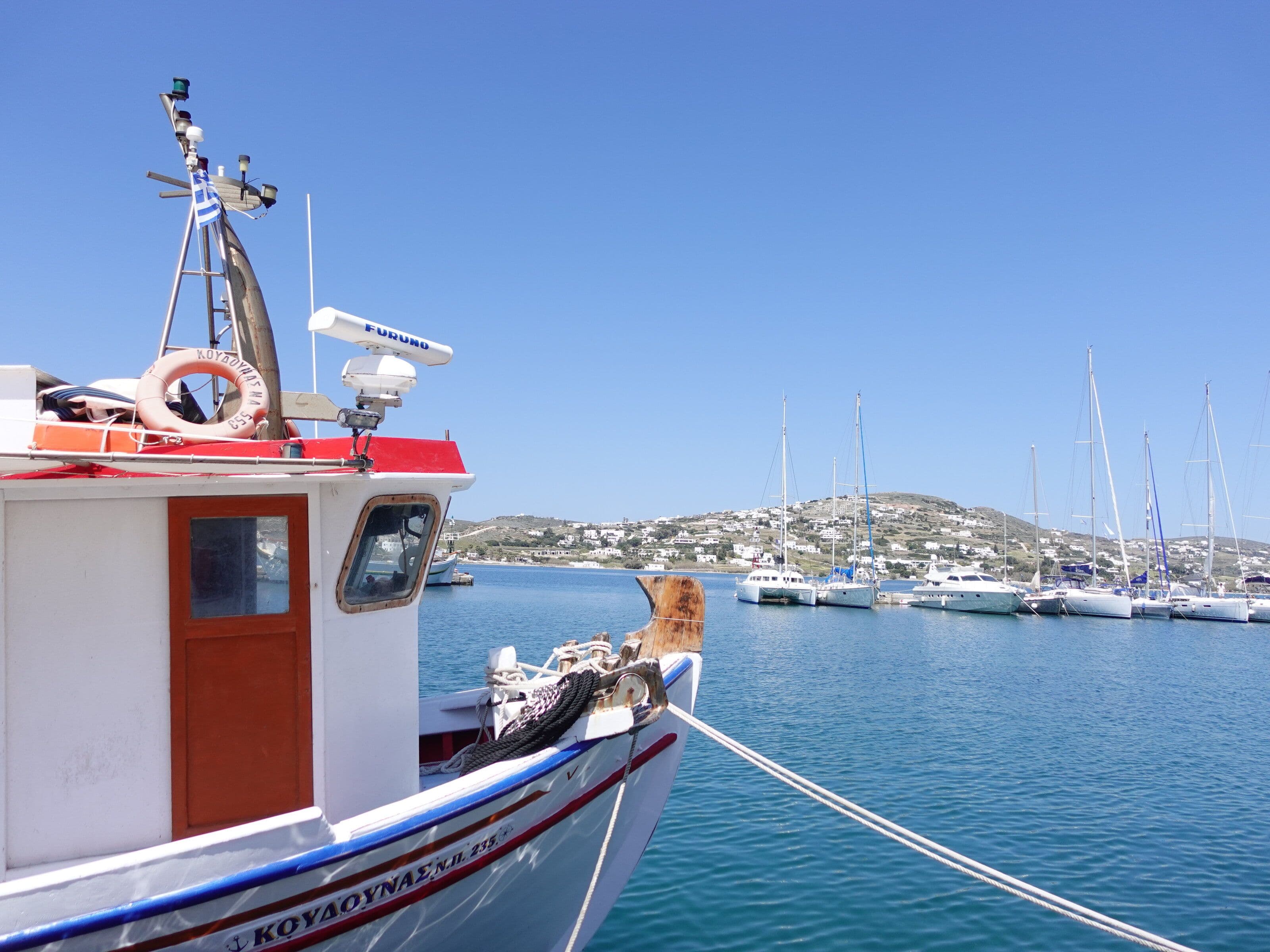A white and red Greek fishing boat named Koulounas moored in a calm blue harbor with sailboats and a hillside village in the background