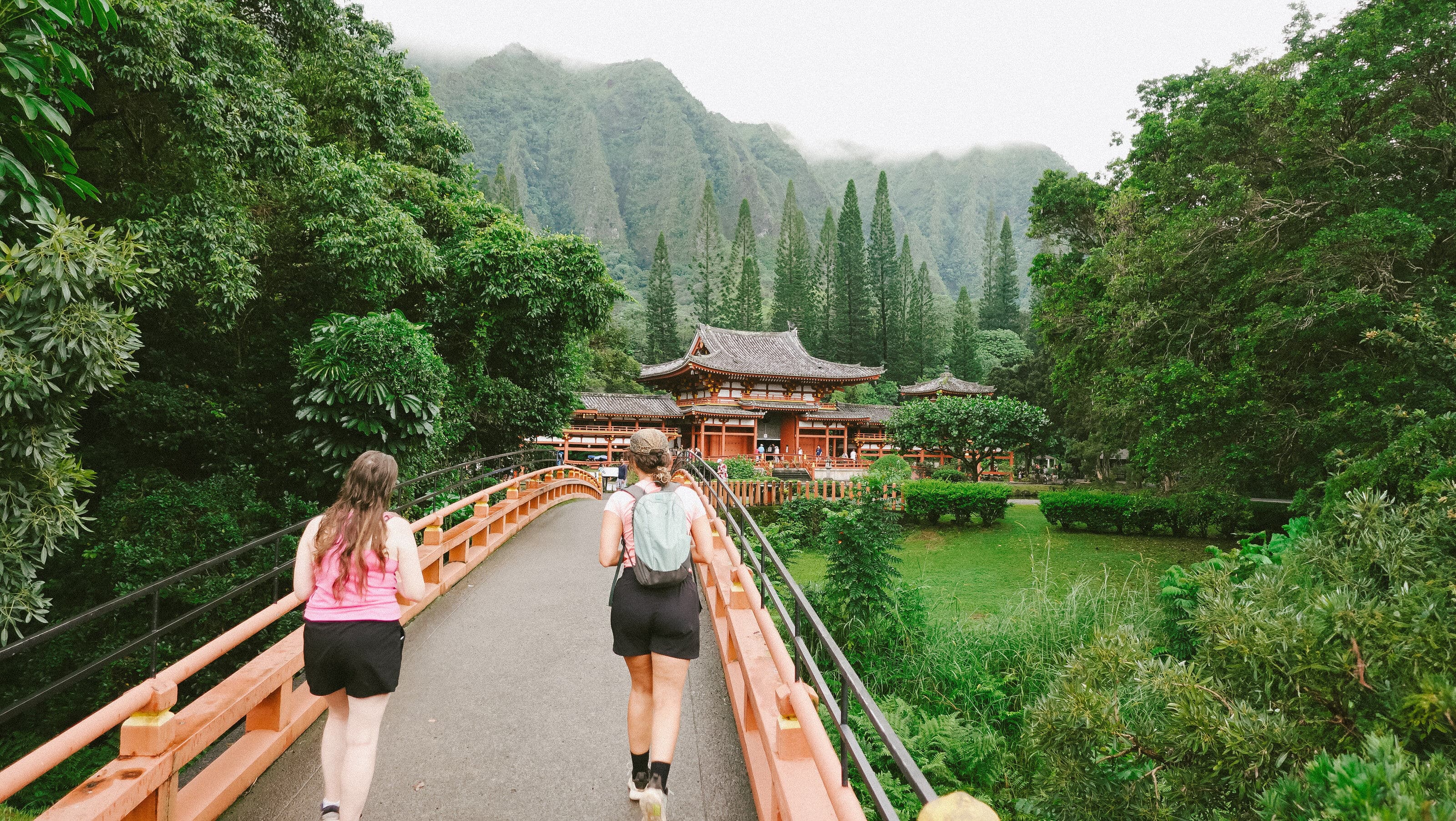 Two women walk along a red bridge path toward the Byodo-In Temple surrounded by lush greenery and dramatic misty mountains in Hawaii
