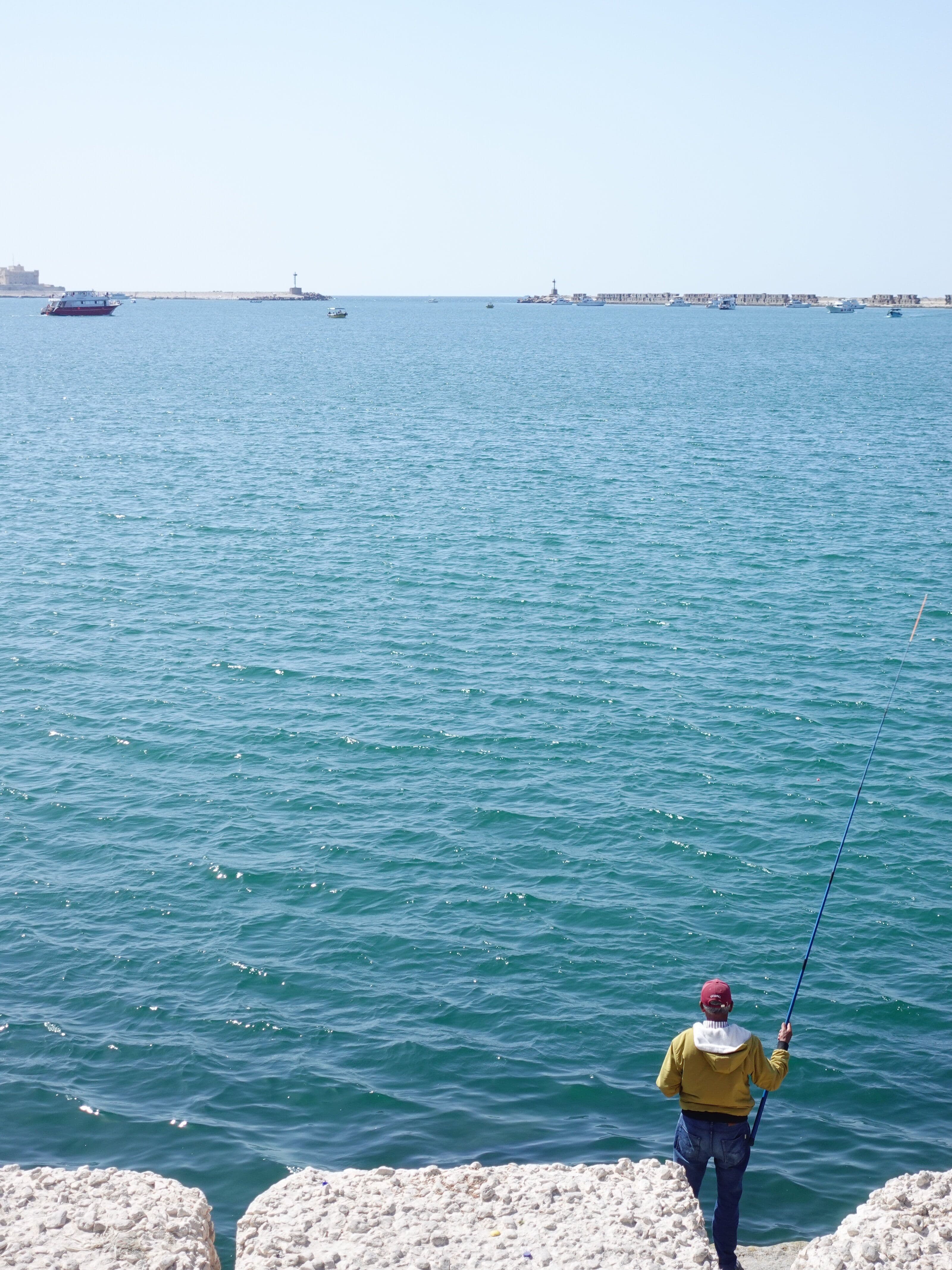 A person in a yellow jacket and red cap fishing with a long rod from a white stone seawall overlooking a calm turquoise harbor with boats in the distance
