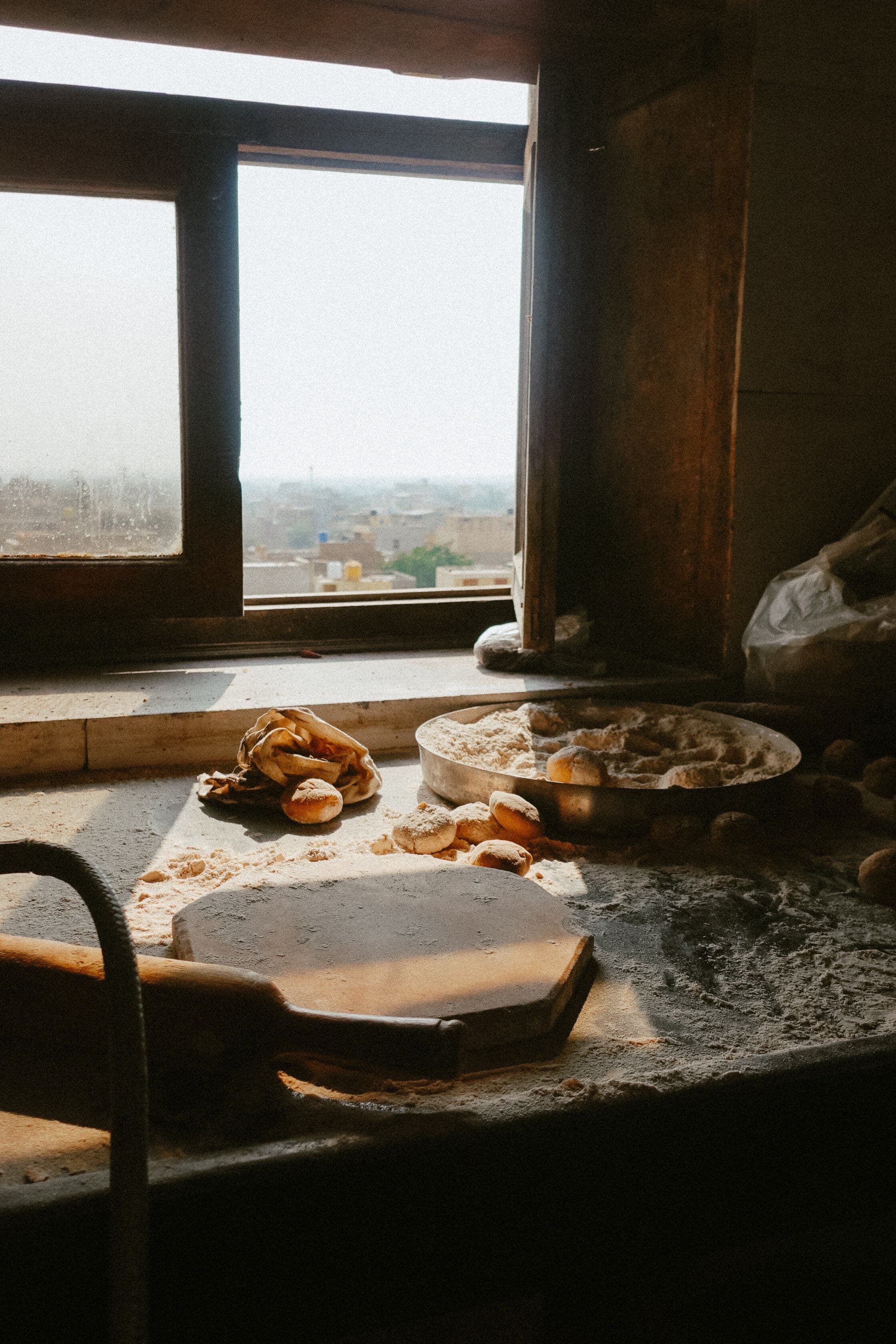 Flour-dusted bread dough balls and a wooden rolling board on a stone counter in a rustic bakery with an open window overlooking a hazy cityscape